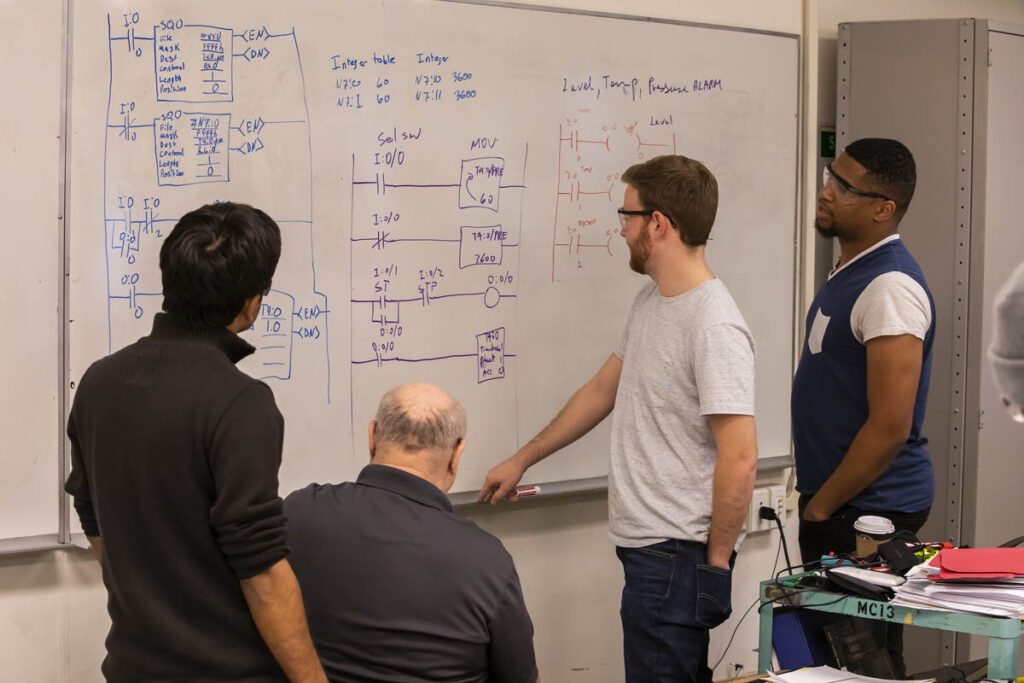 Students and instructor looking at a whiteboard with some schematics drawn on it