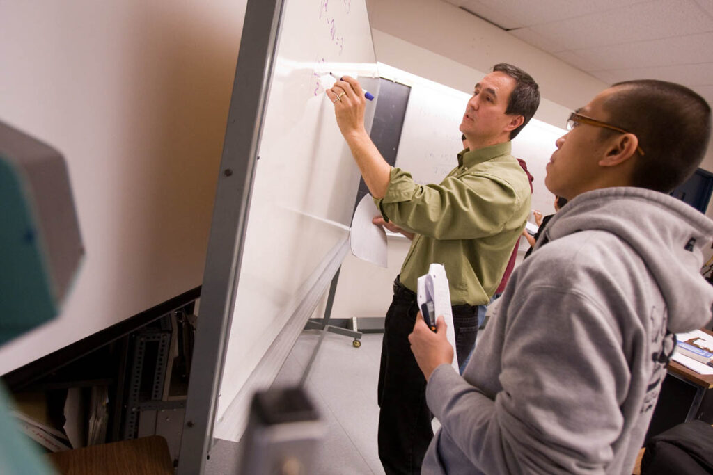 Instructor writing on white board while student talks to him