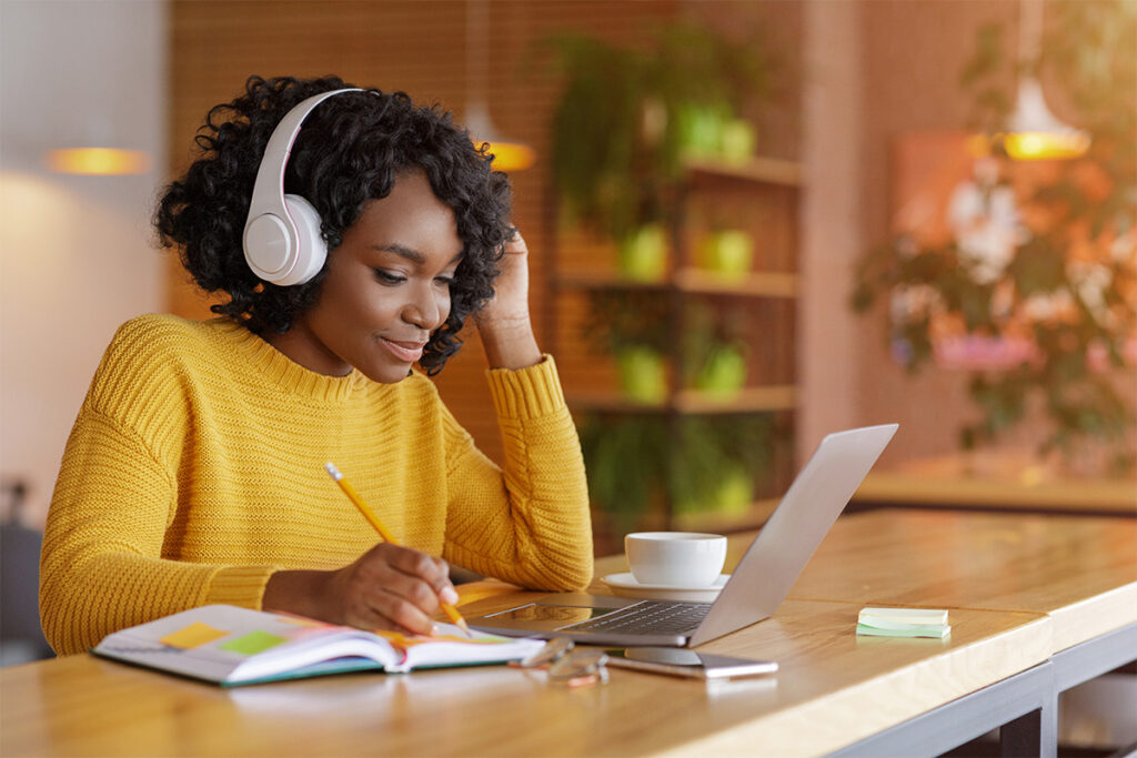 Woman wearing headphones looking at a textbook and laptop while taking notes