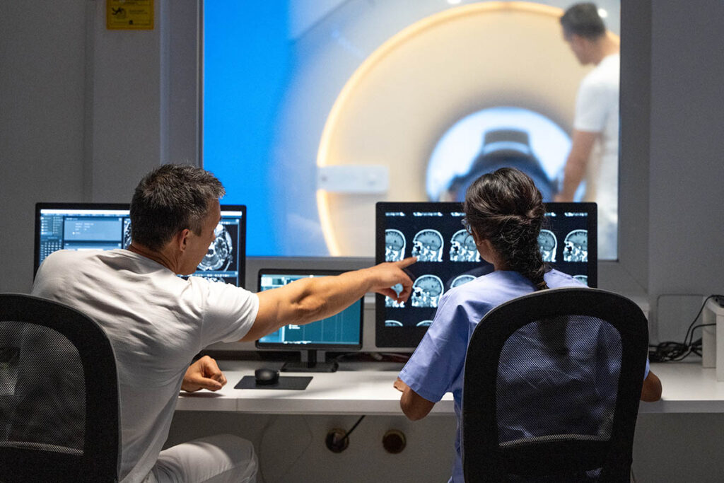 Two medical professionals looking at MRI scans while a technician slides a patient into the scanner in the background