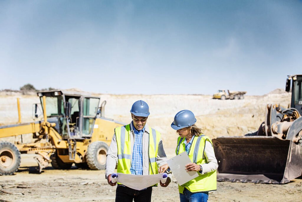 Two people in hard hats and safety vests looking at blueprints at a construction site