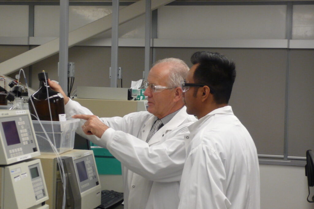 Two men in white coats and safety glasses talking in a lab