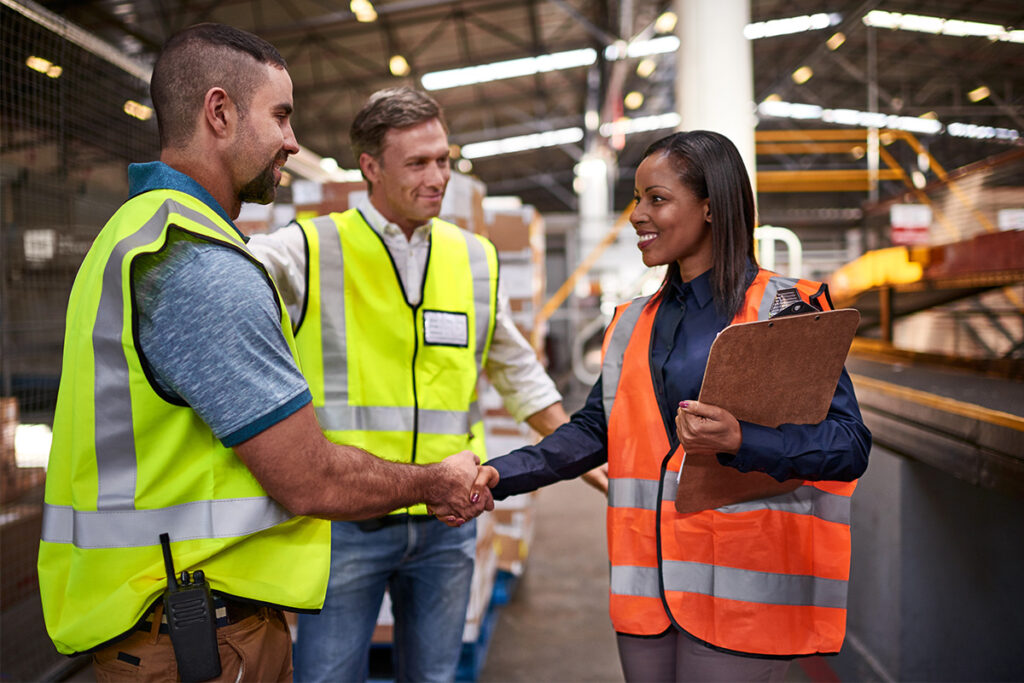 Three people in safety vests shaking hands in a distribution centre 