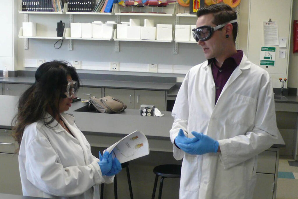 Two people talking in a lab wearing white coats and safety glasses