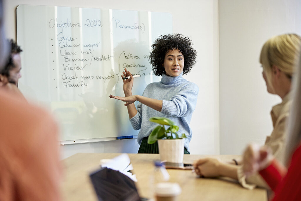 Woman standing at front of room beside whiteboard and talking to others in the room