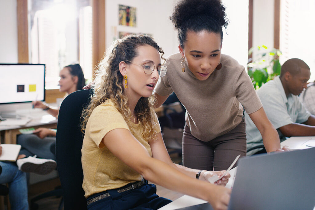 Two women looking at a laptop screen in a busy office