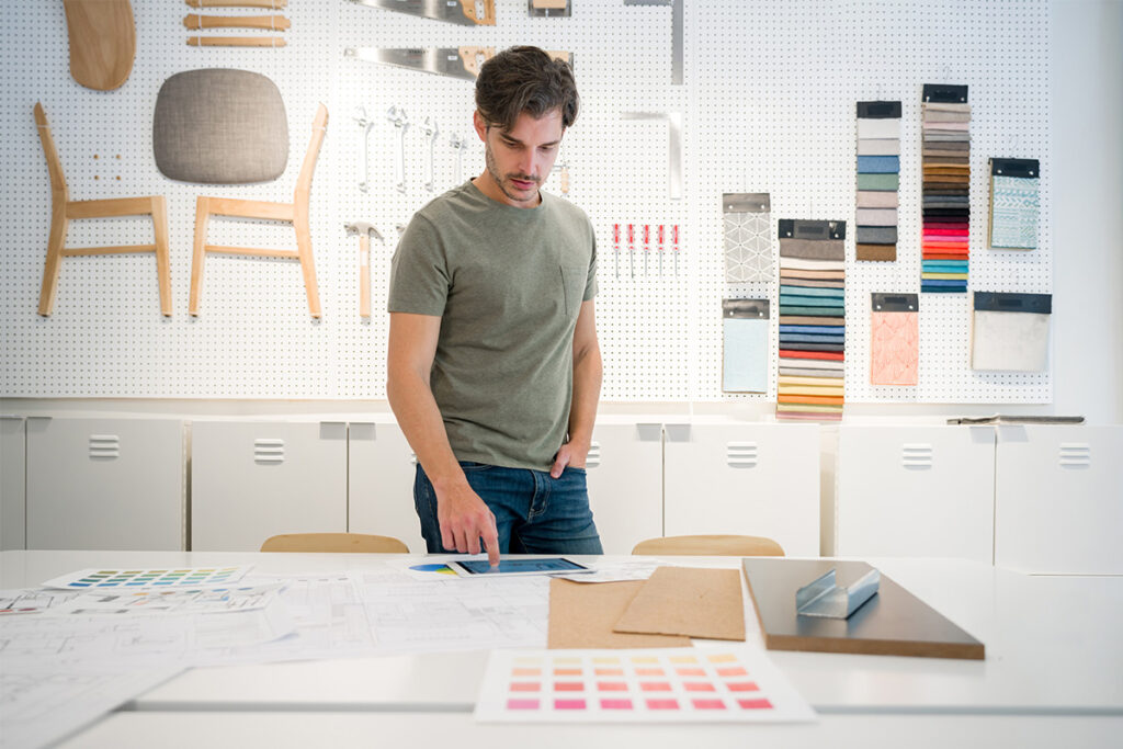 Man standing in design studio with colour swatches and a deconstructed chair on the wall, and is looking at papers on a table