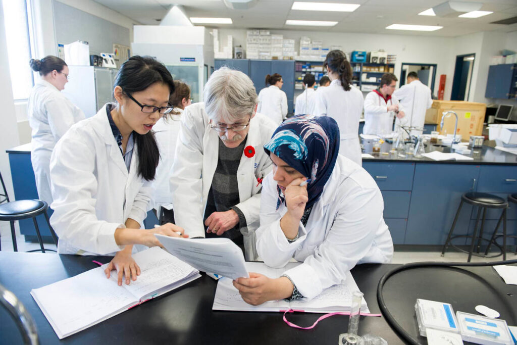 Students wearing white lab coats reviewing notes with their instructor in a science laboratory