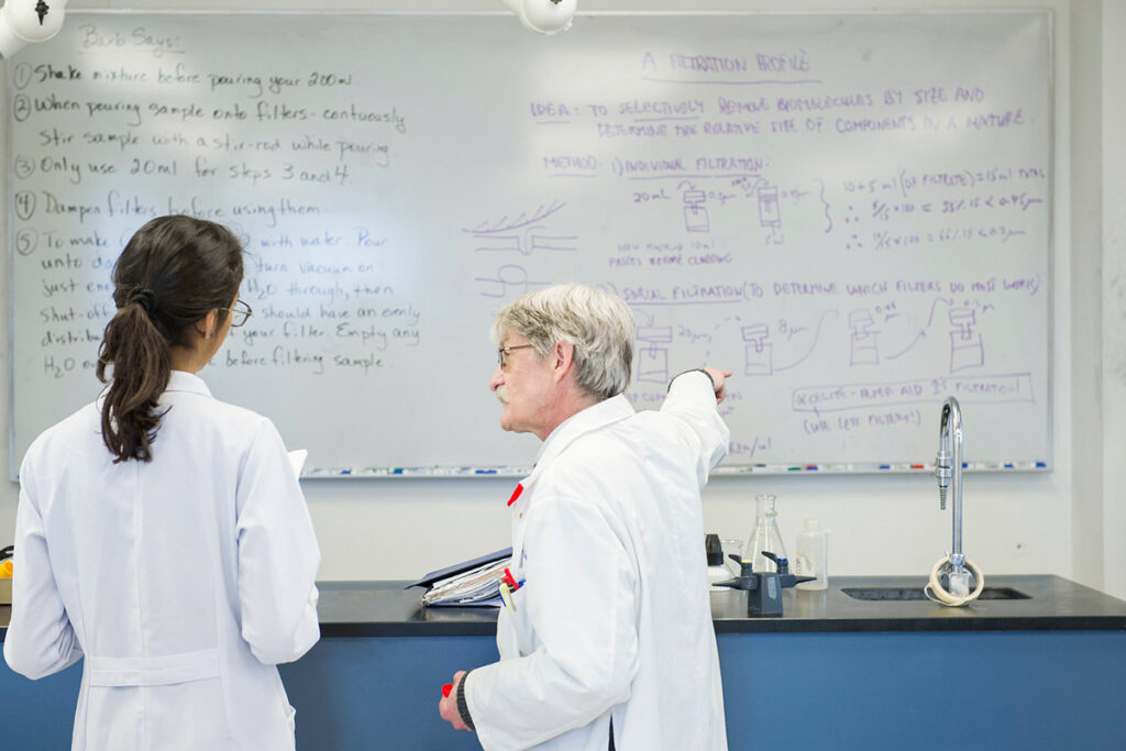 Instructor and student in white lab coats looking at a whiteboard with many notes written on it