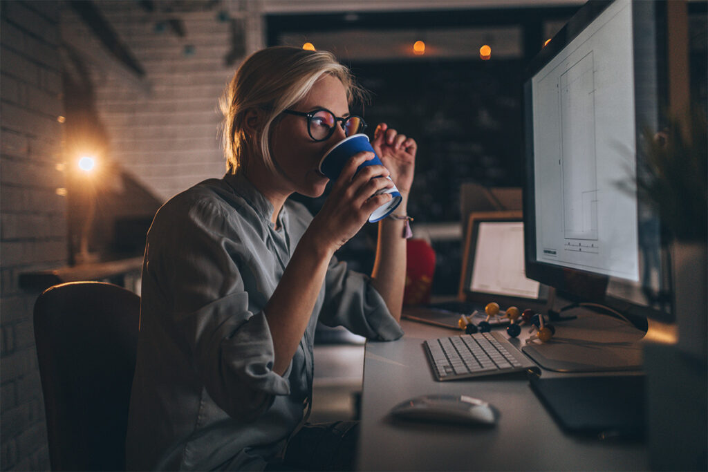 Woman drinking something while working at a computer in a dimly lit office