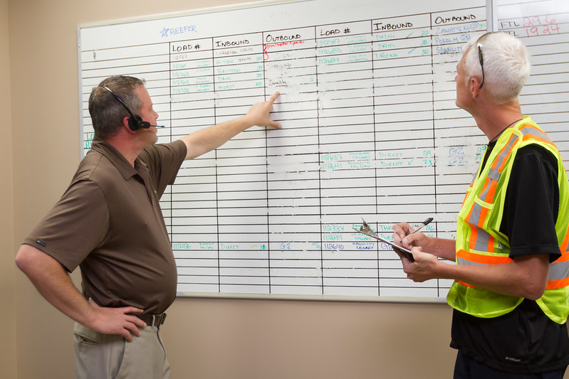 Two men looking at a whiteboard with logistics written on it