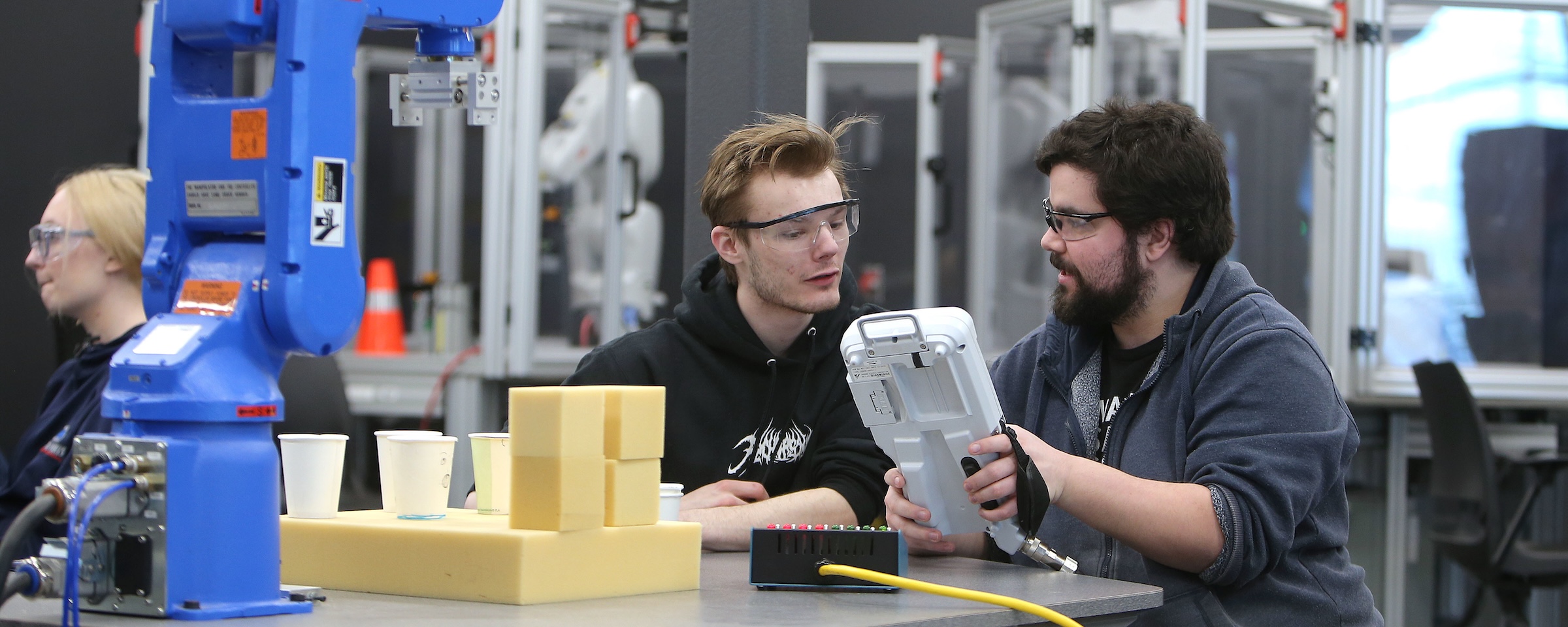 Two students wearing safety glasses work with a handheld industrial robot controller beside robotic manufacturing equipment in a lab.