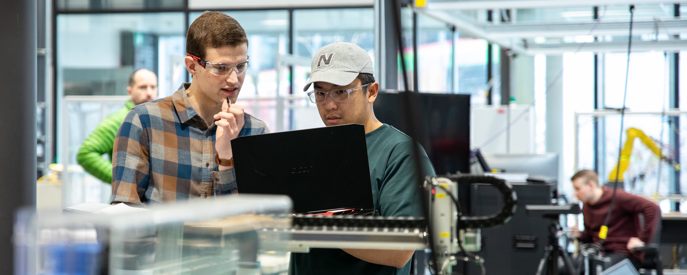 Two students wearing safety glasses work together on a laptop beside automated manufacturing equipment in a mechatronics lab.