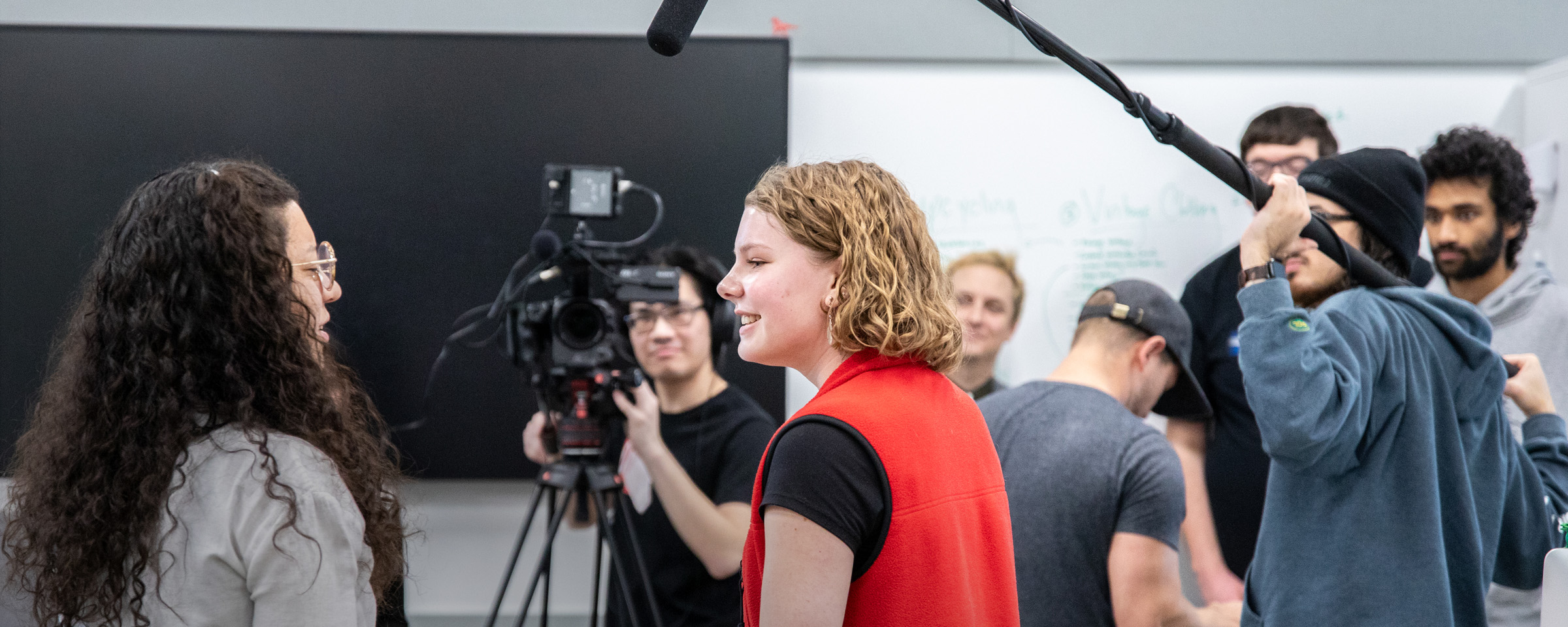 Students in a film production class work collaboratively on set, with one student operating a boom mic, another behind a video camera, and two students engaged in an on-camera interview, surrounded by classmates in a studio setting.