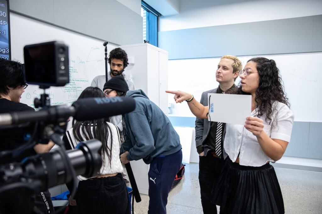 Student operating professional film camera on a studio set, adjusting camera focus under bright production lights.