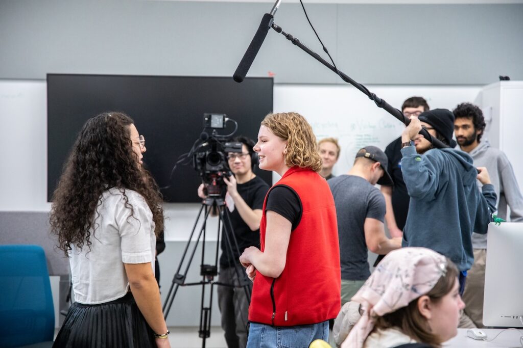 Group of students directing a scene on a soundstage, with lighting rigs and cameras visible in the background.