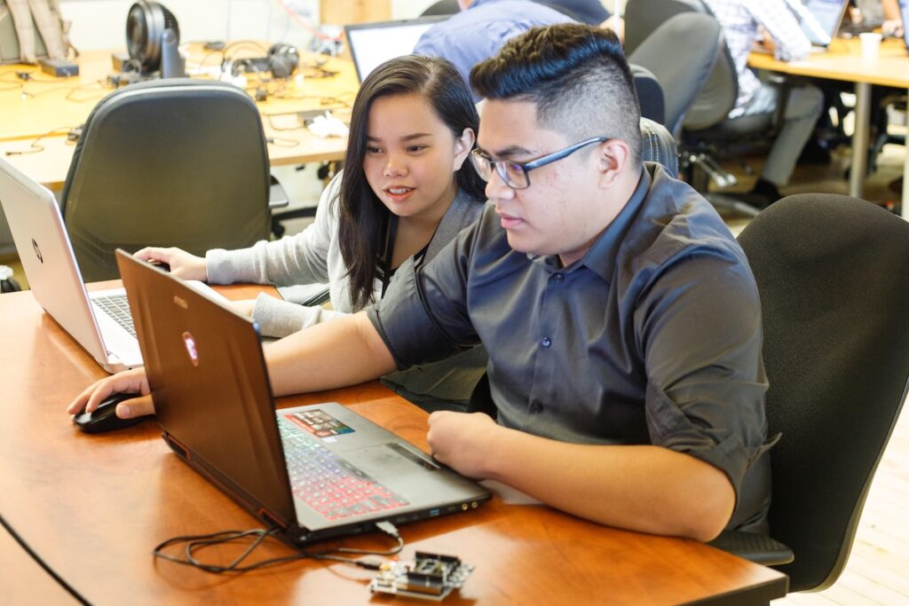 Two students seated at long tables in a collaborative workspace, each working on laptops