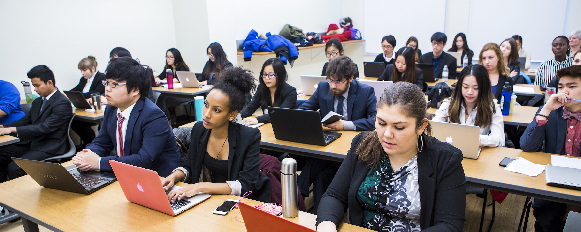 Business students in professional attire attending a lecture in a large classroom with laptops