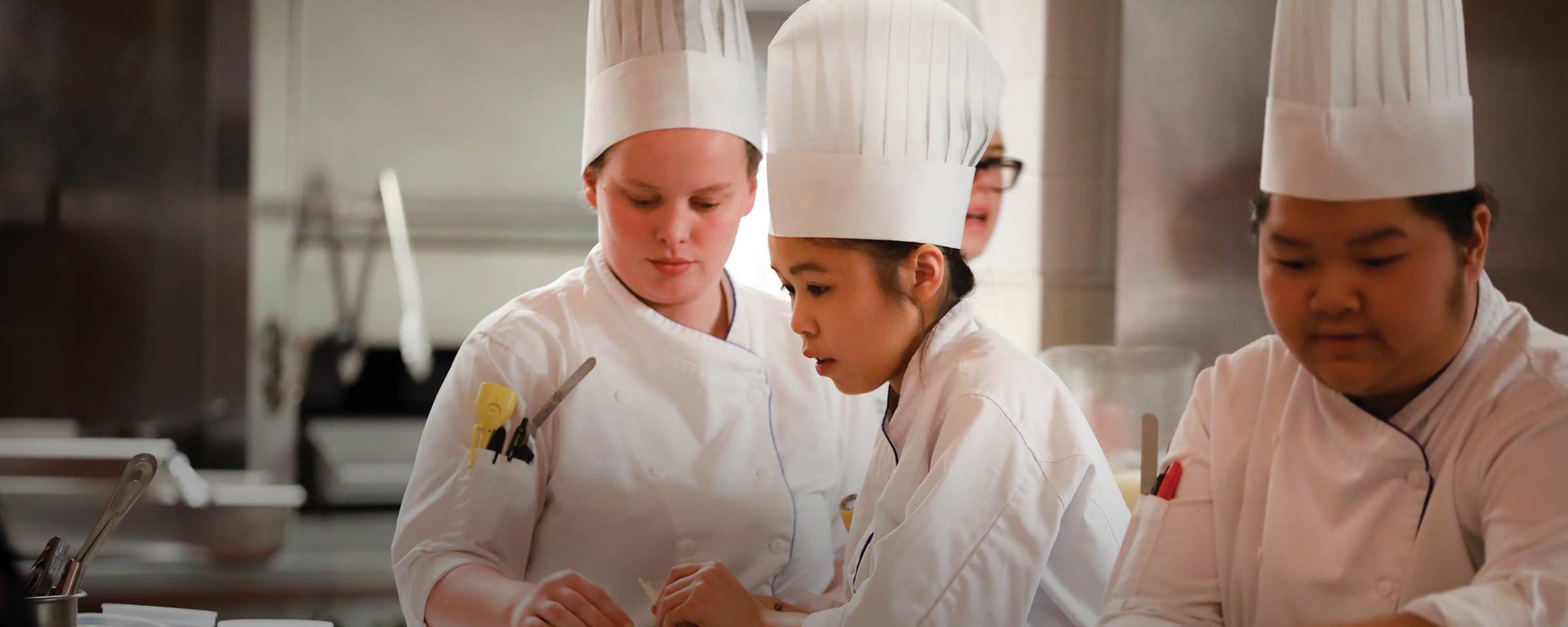 Three culinary students in white chef uniforms and tall toques collaborate over a workstation in a professional kitchen.