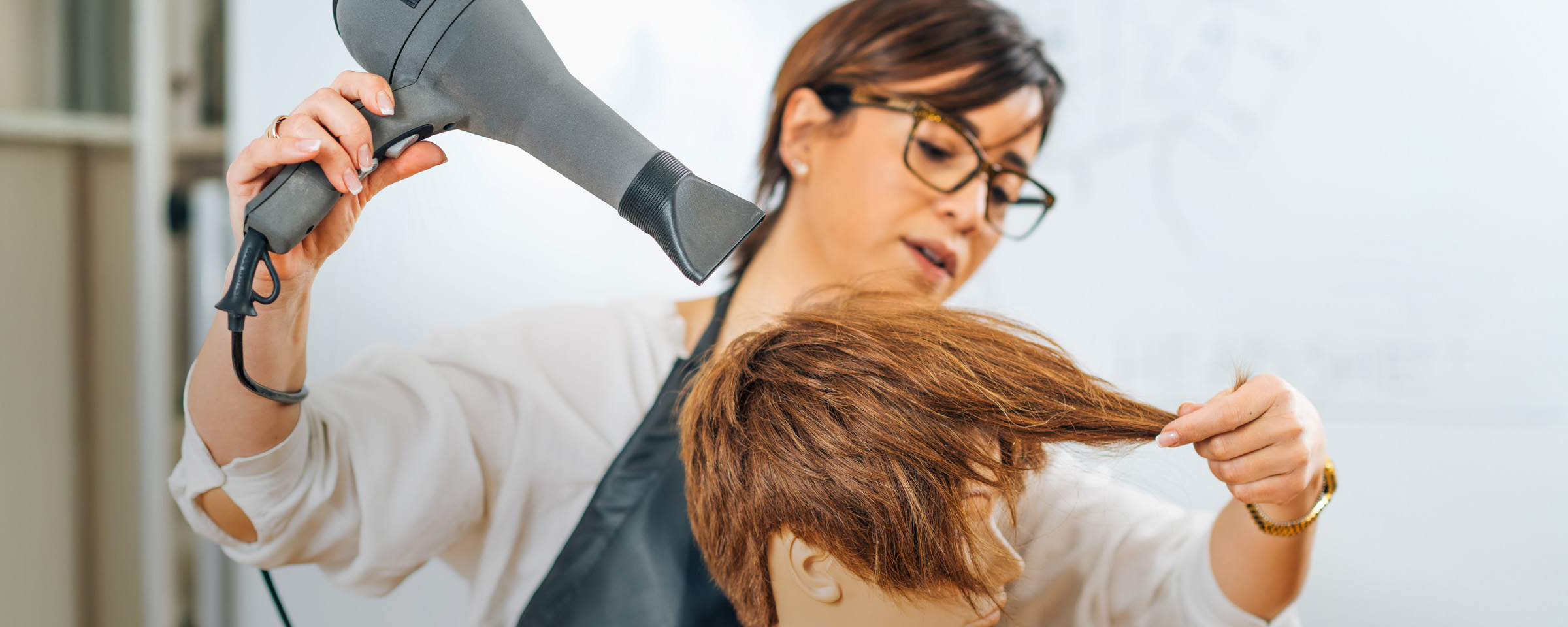 A hairstyling instructor wearing glasses uses a blow dryer and her hands to style a client's damp auburn hair in a salon setting.