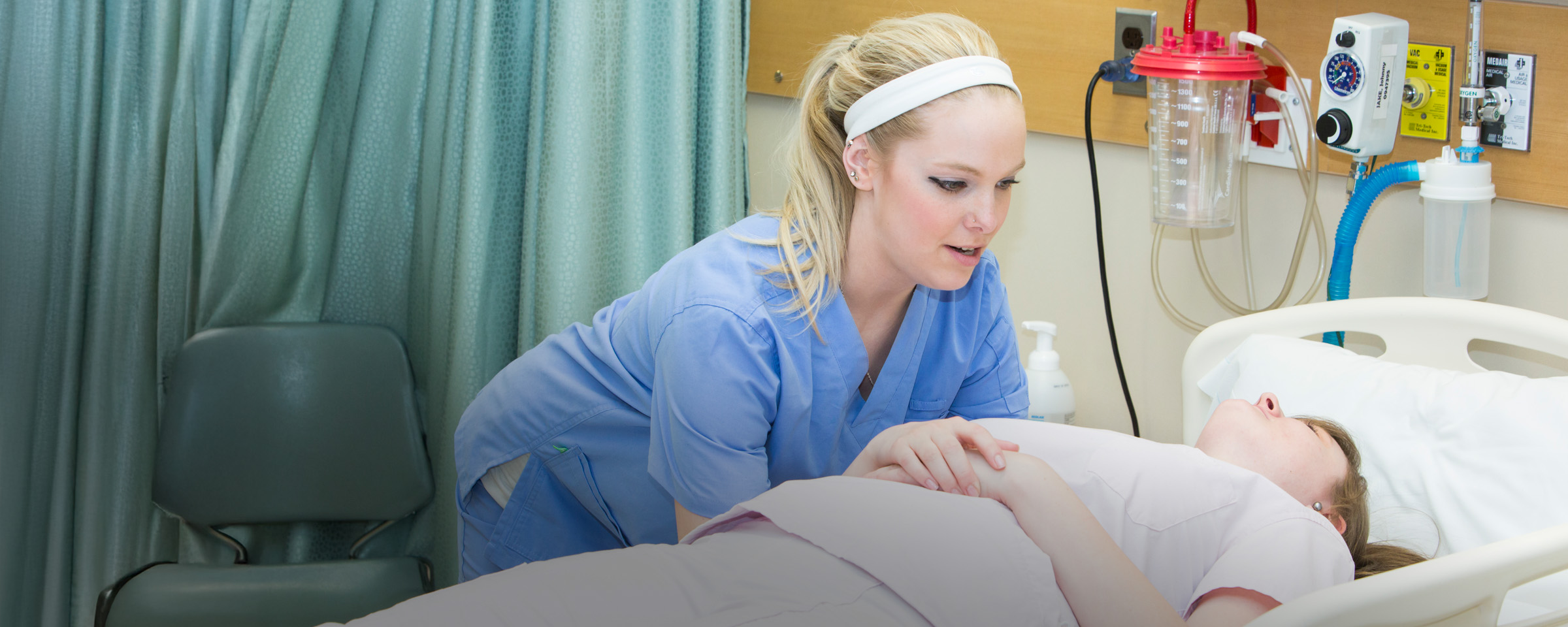 A nursing student in blue scrubs leans over a patient lying in a hospital bed in a clinical simulation room.