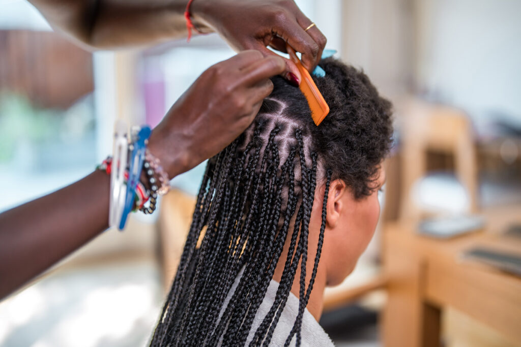 Close-up of a hairstylist using an orange comb to section a client's hair while installing box braids in a salon setting.
