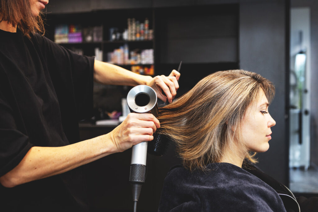 A hairstylist in a black top blow-dries a seated client's highlighted bob-length hair in a modern salon with product shelves in the background.