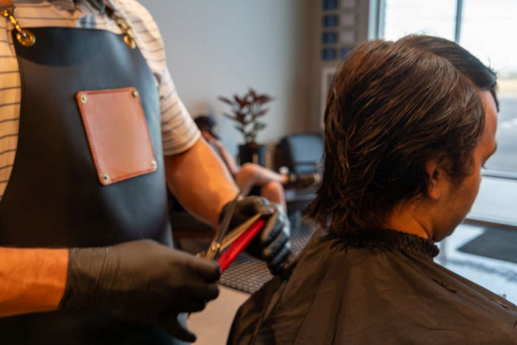A barber wearing a leather apron and black gloves uses clippers on a male client's damp dark hair in a barbershop.