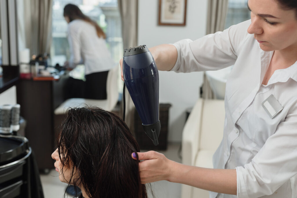 A hairstylist in a white coat blow-dries a client's long dark wet hair in a salon, with a mirror reflecting another stylist in the background.