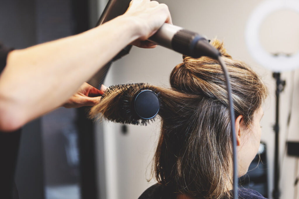Close-up of a hairstylist using a round brush and blow dryer to style the back section of a client's medium-length brunette hair.