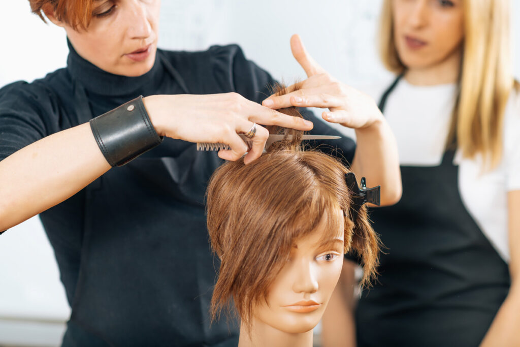 A hairstyling instructor demonstrates a scissor cut on a mannequin head while a student in a black apron observes closely.