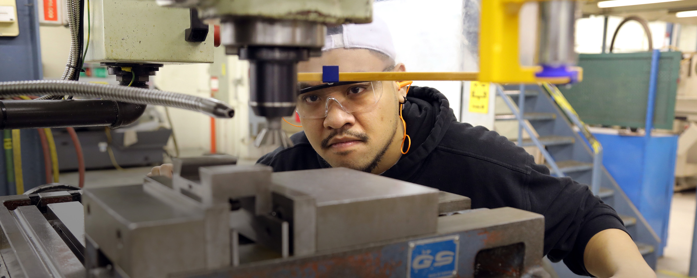 A male student wearing safety glasses peers closely at a milling machine, carefully aligning a metal workpiece in an industrial workshop.