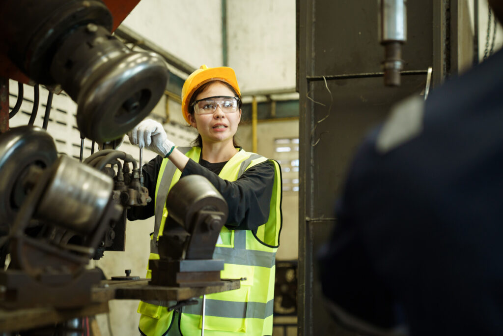 A female millwright student wearing a yellow hard hat, safety glasses, and high-visibility vest operates industrial machinery in a manufacturing facility.
