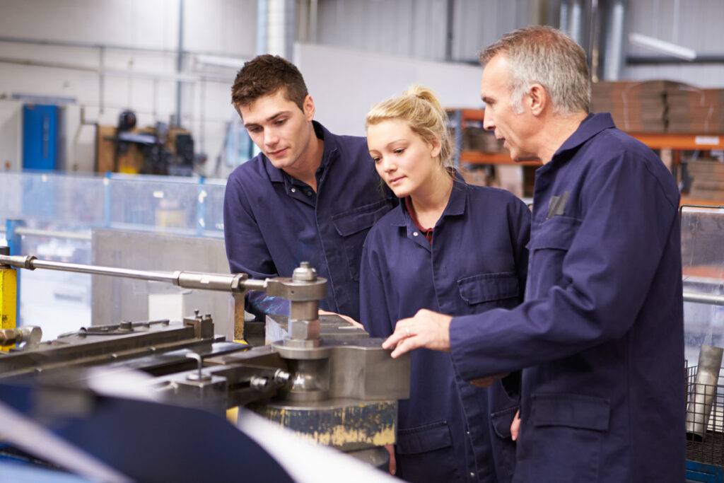 A senior millwright instructor in navy coveralls supervises two apprentices as they examine a metal tube bending machine on the shop floor.
