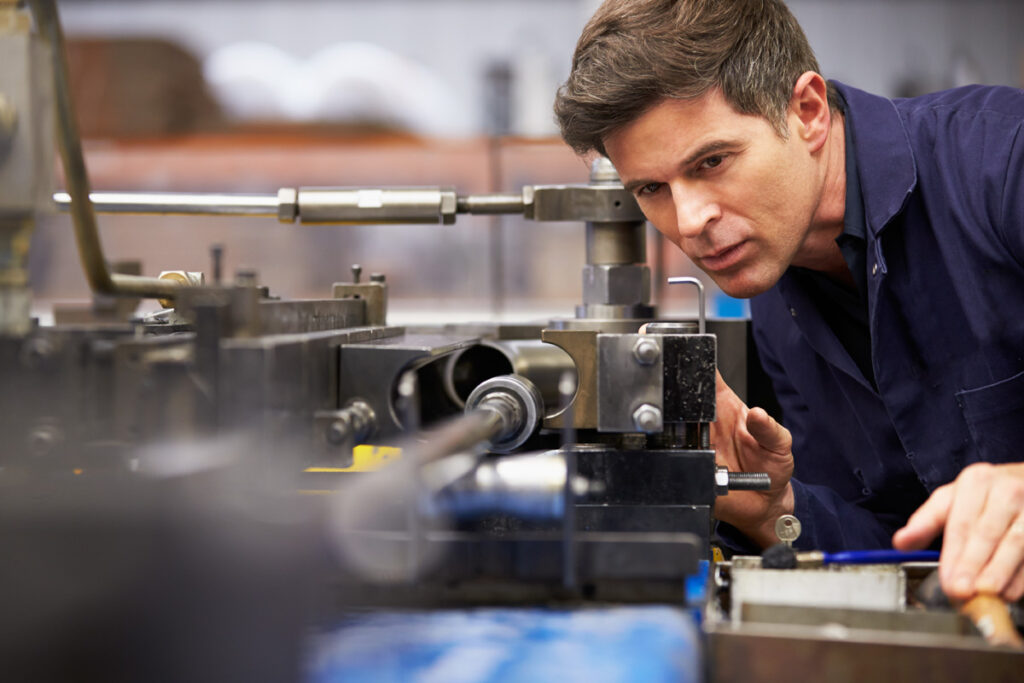 A male millwright in navy coveralls peers intently at the components of a lathe machine in an industrial workshop.