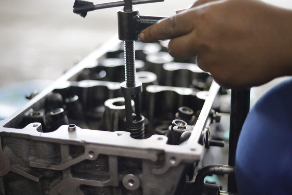 Close-up of a technician's hands using a valve spring compressor tool on a disassembled engine cylinder head.