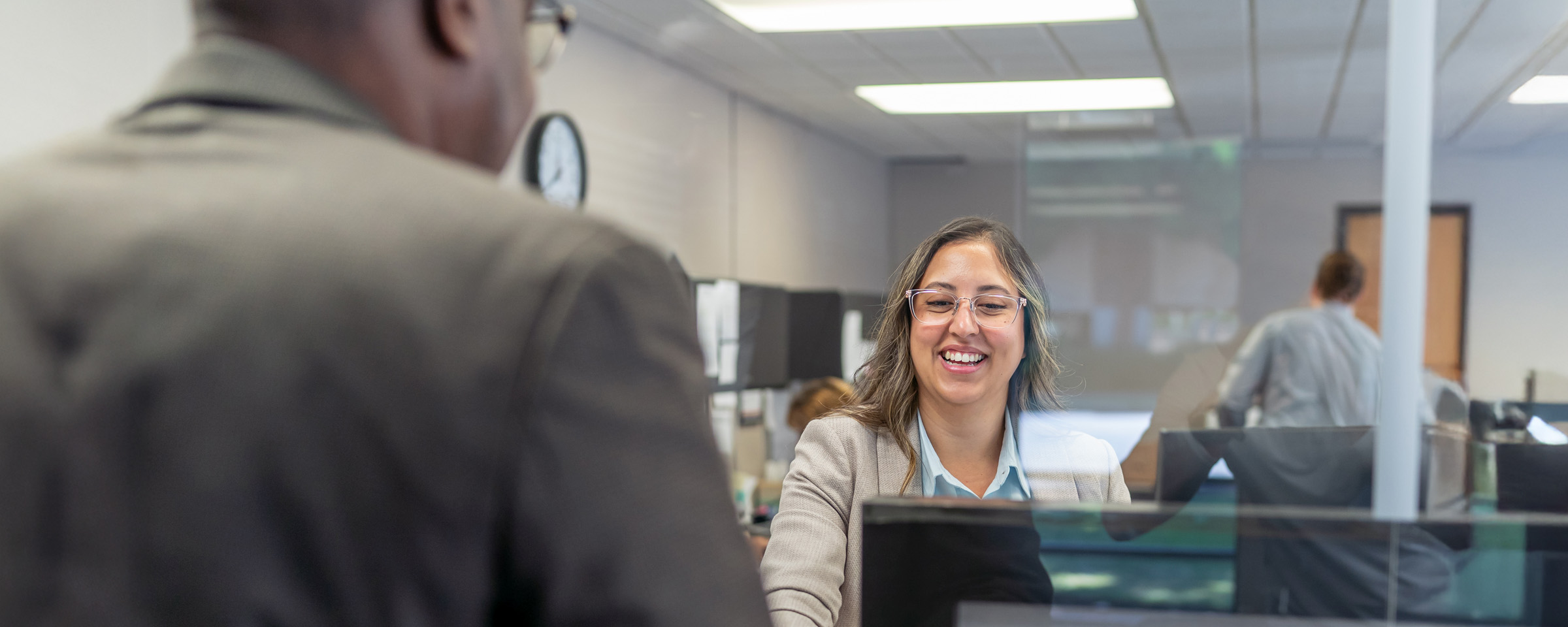 A smiling female medical office assistant in professional attire speaks with a patient across a reception desk in a healthcare administration office.