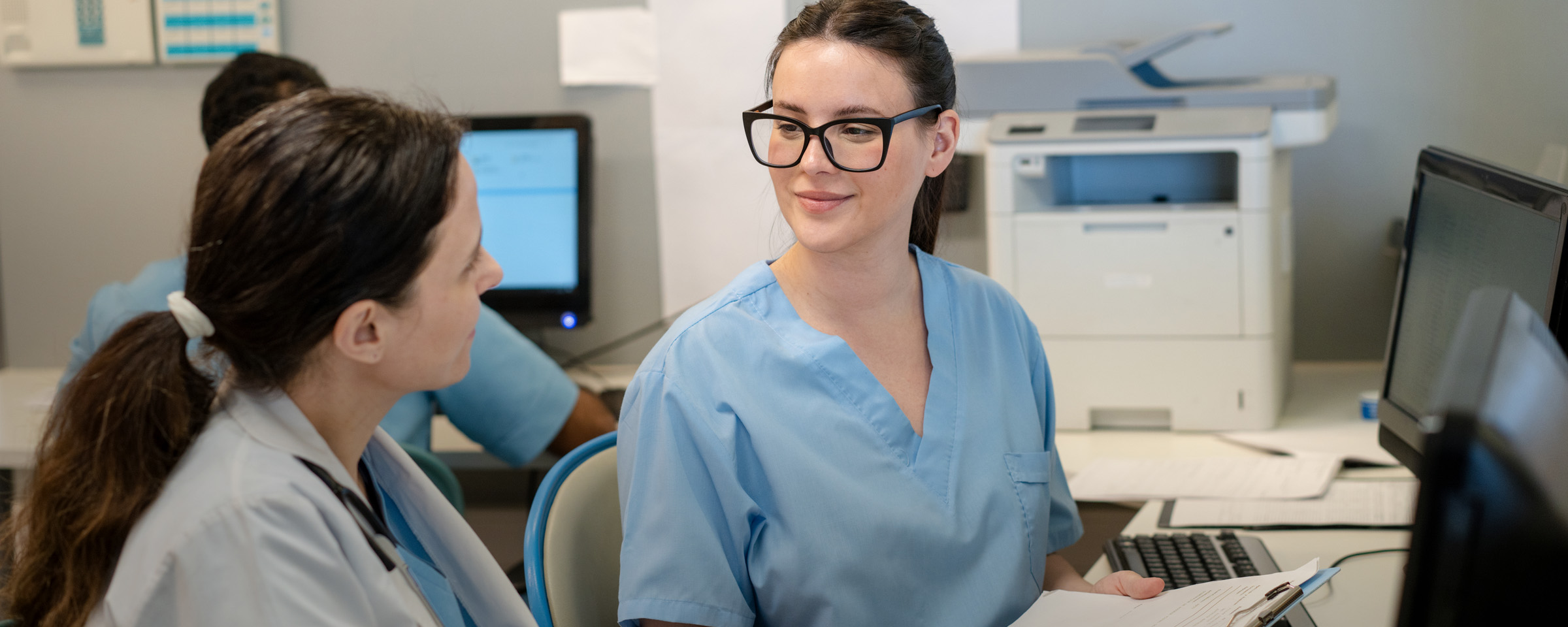 Two healthcare students in scrubs sit together at a workstation, reviewing a clipboard and conversing in a medical office setting.