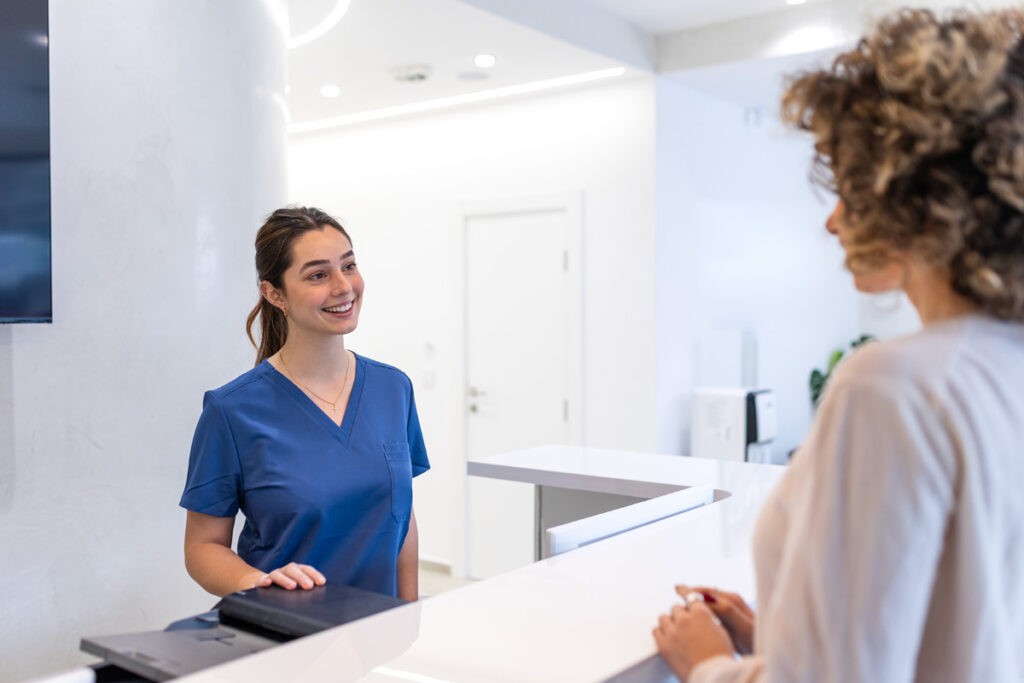 A smiling medical assistant in blue scrubs greets a curly-haired patient across a clean white reception desk in a modern clinic.