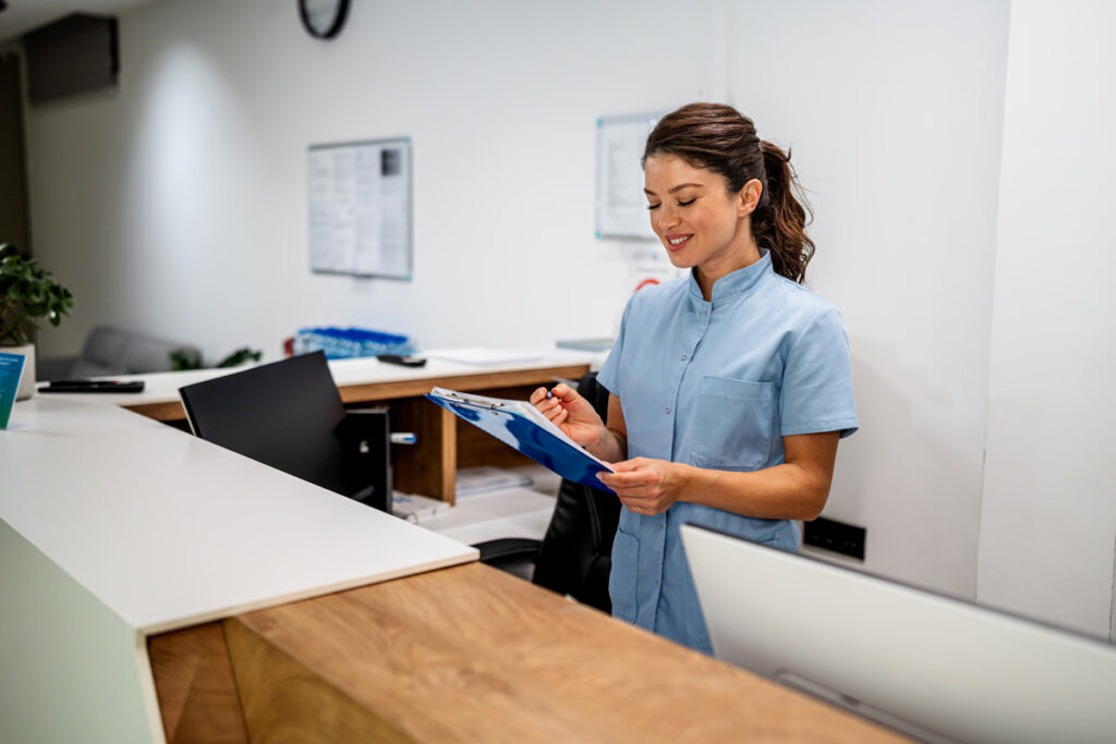 A medical assistant in light blue scrubs reviews a clipboard while standing at a reception desk in a medical office.