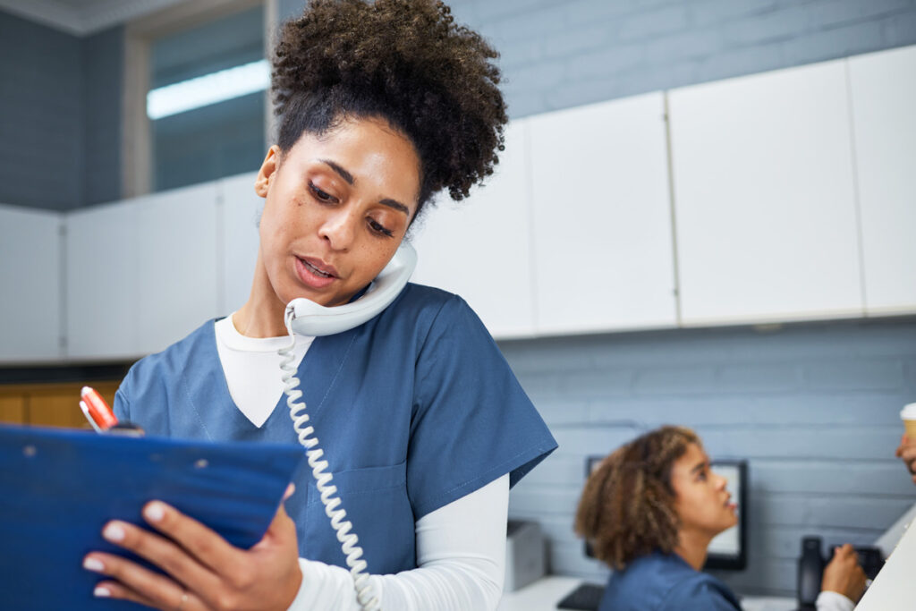 A medical assistant in navy scrubs holds a corded phone to her shoulder while reviewing a clipboard, with a colleague working at a computer in the background.
