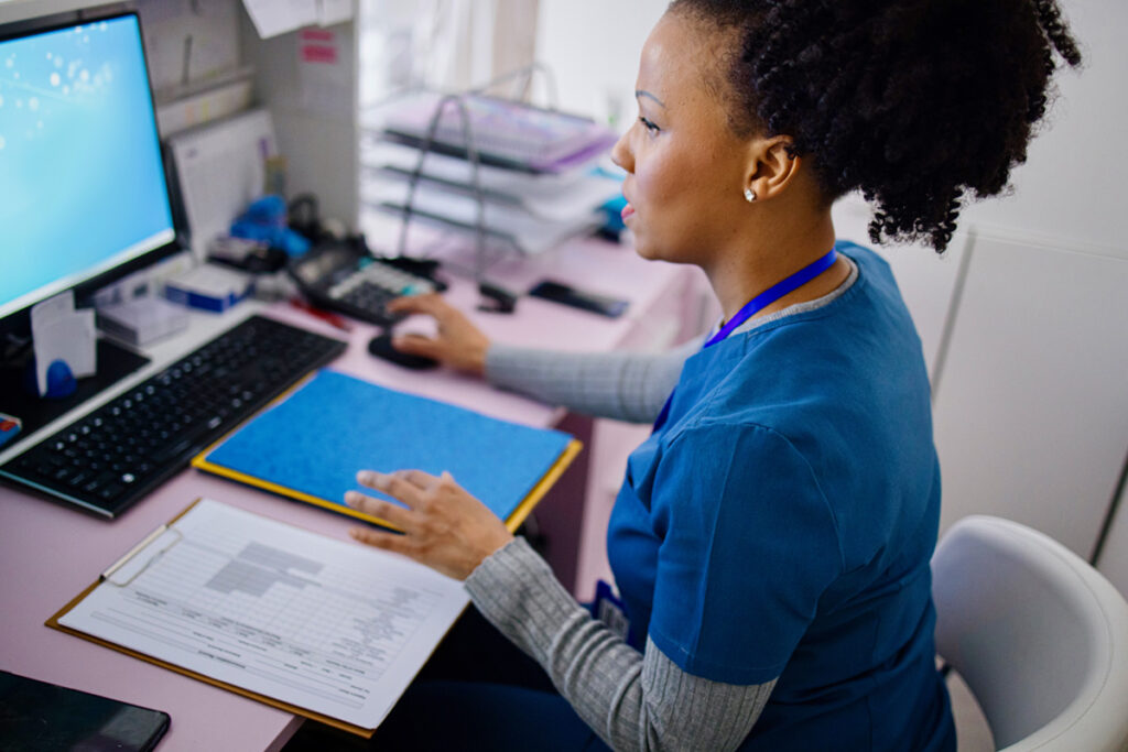 A medical office administrator in blue scrubs and a lanyard uses a computer mouse while referencing patient forms at a healthcare workstation.