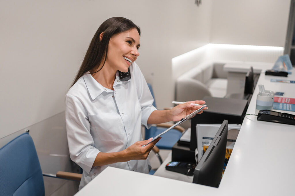 A smiling medical receptionist in a white blouse talks on a corded phone while reviewing a tablet with a stylus at a clinic front desk.