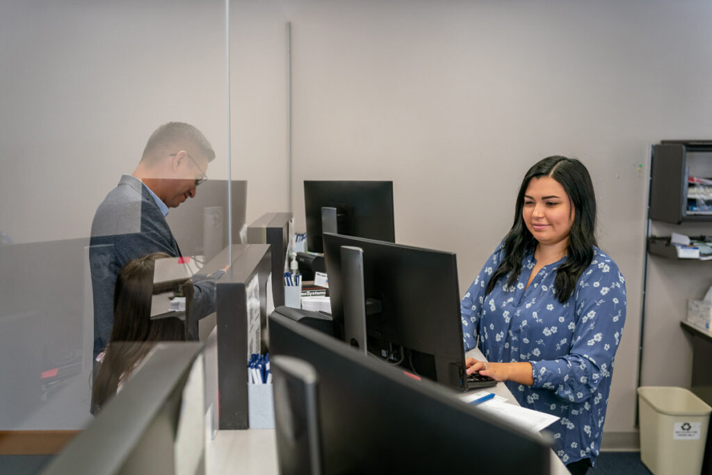 A medical assistant in a blue floral blouse works at a computer workstation behind a reception desk, with a colleague and patient visible in the background.