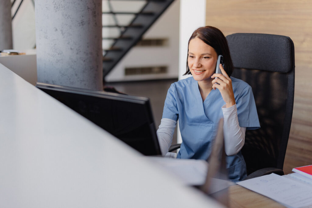 A smiling medical assistant in blue scrubs talks on a phone while working at a computer at a healthcare reception desk.