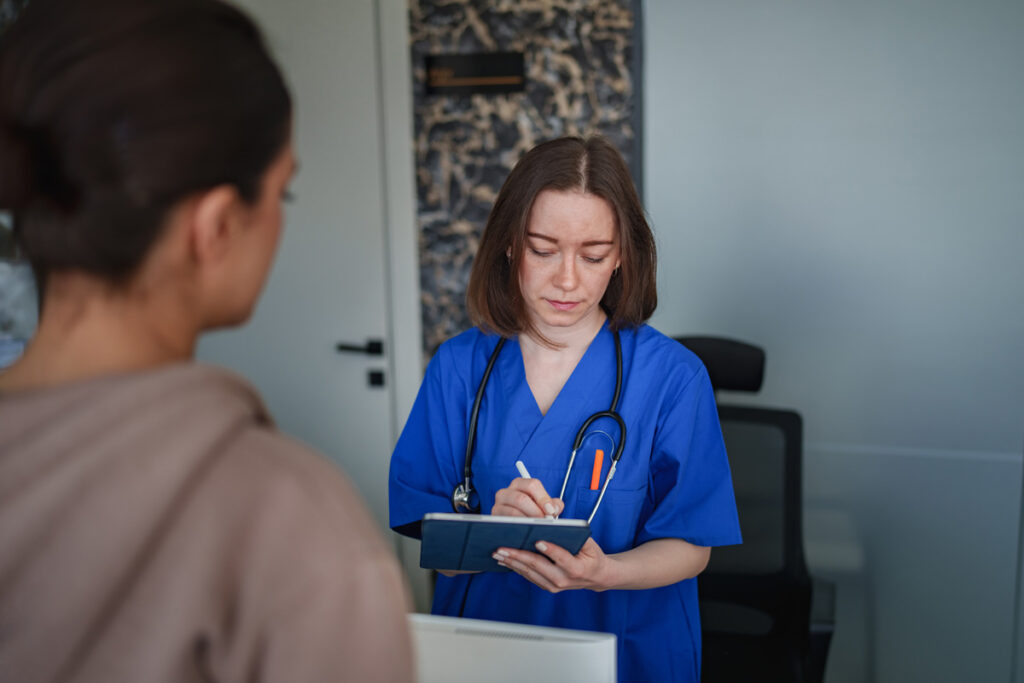 A medical assistant in blue scrubs with a stethoscope writes notes on a clipboard while consulting with a patient in a clinical office.