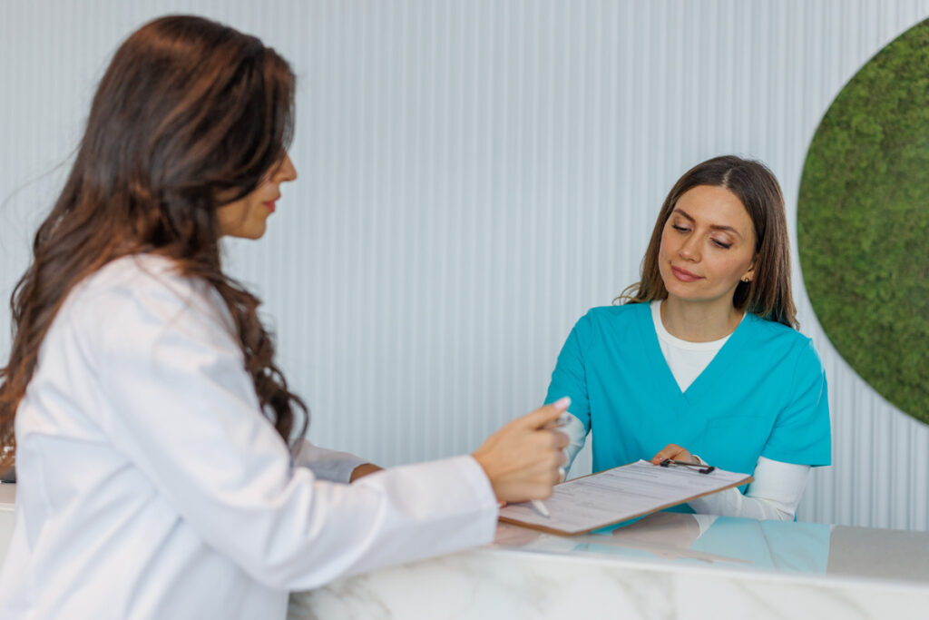 A medical assistant in teal scrubs reviews paperwork on a clipboard with a physician in a white coat at a clinic reception desk.