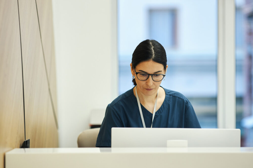 A medical office administrator wearing glasses and dark scrubs focuses on a laptop at a reception desk in a healthcare facility.