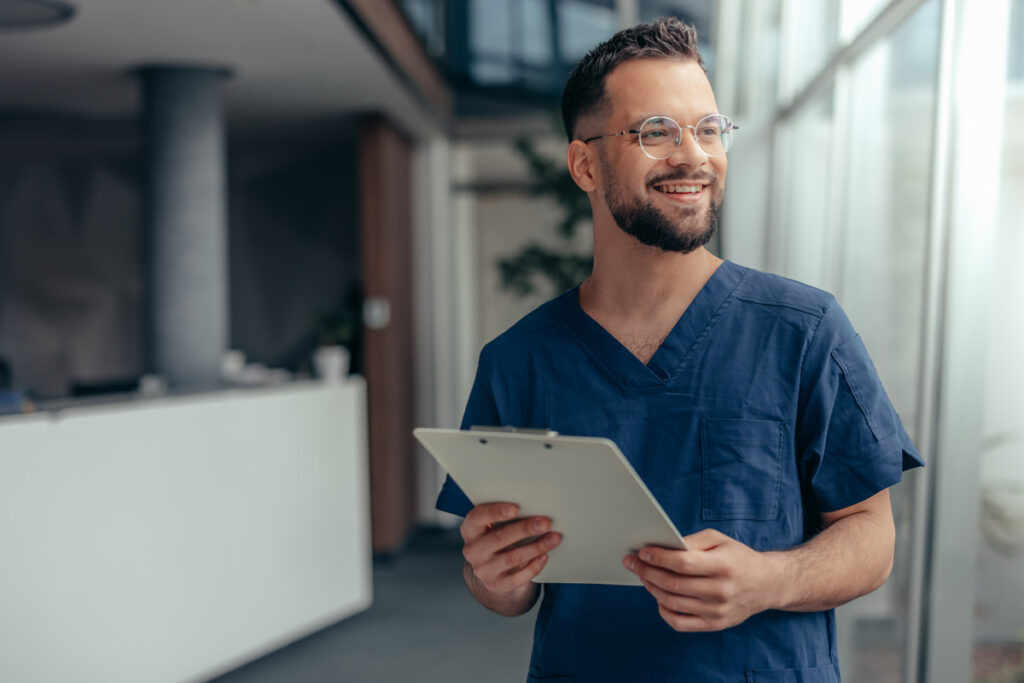 A smiling male healthcare professional in navy scrubs and glasses holds a clipboard in a modern medical office corridor.