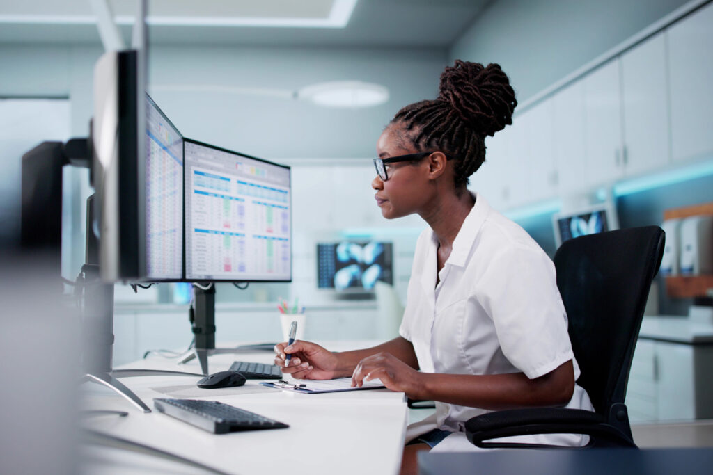 A female healthcare worker in a white lab coat and glasses reviews patient data on dual monitors in a clinical workstation environment.